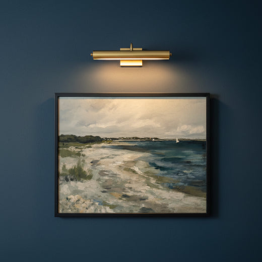 A framed landscape painting depicting a sandy beach meeting the blue water of the sea, with a cloudy sky and a small sailboat on the horizon, presented in a black frame.