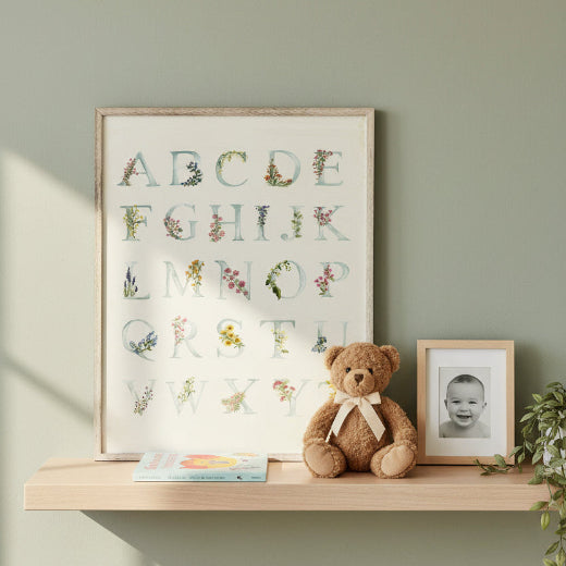 A framed alphabet art print featuring the letters A through Z styled with delicate watercolor floral and botanical illustrations. The print has a light cream background with pale blue-gray lettering.