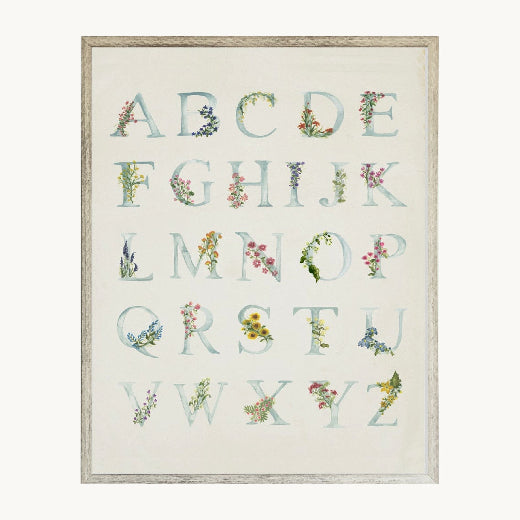 A framed alphabet art print featuring the letters A through Z styled with delicate watercolor floral and botanical illustrations. The print has a light cream background with pale blue-gray lettering.