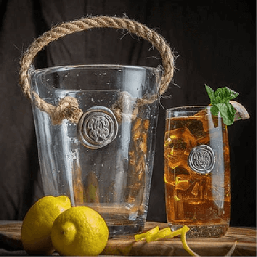 Glass ice Bucket with a pewter intiial emblem and rope handle on a wooden surface with a lemon placed beside it.