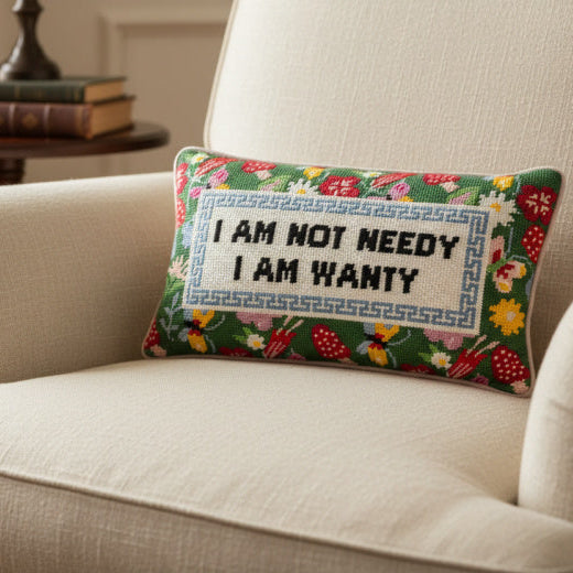 Needlepoint pillow with the phrase I am not needy I am wanty on a beige armchair in a home setting