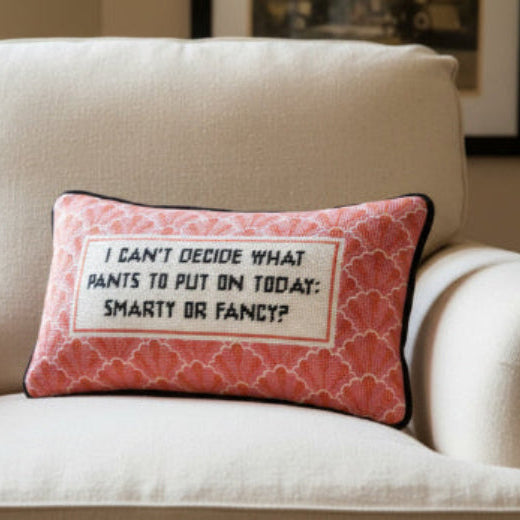 Decorative needlepoint pillow with I can't decide what pants to put on today: smarty or fancy text sitting on a beige armchair