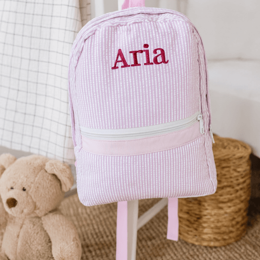 Pink backpack with 'Aria' embroidery, teddy bear, and wicker basket in the background