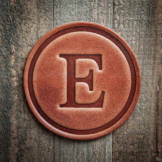 Signature leather initial coaster embossed with the letter E on a rustic wood surface — The Red Hound Gifts