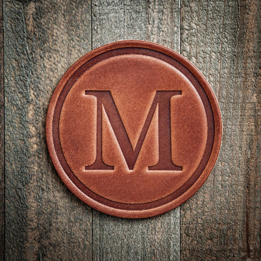 Signature leather initial coaster embossed with the letter M on a rustic wood surface — The Red Hound Gifts