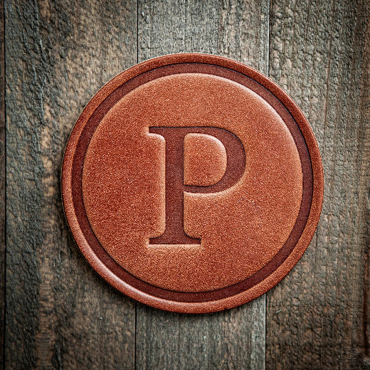 Signature leather initial coaster embossed with the letter P on a rustic wood surface — The Red Hound Gifts