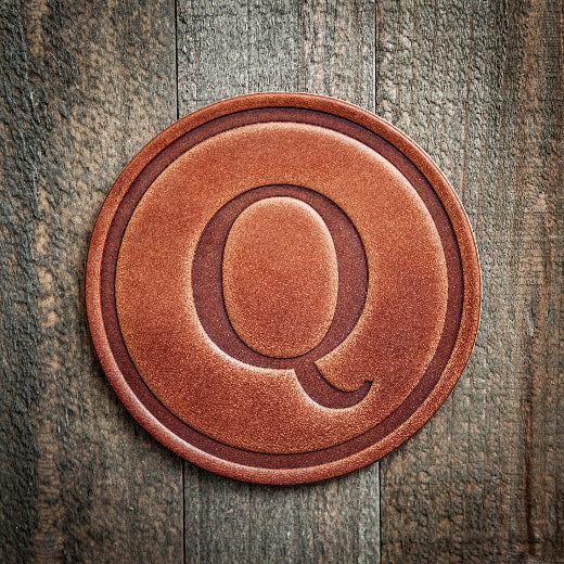Signature leather initial coaster embossed with the letter Q on a rustic wood surface — The Red Hound Gifts