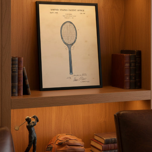 A framed vintage tennis patent print featuring a blue technical drawing of a tennis racket on an aged beige paper background, enclosed in a black frame.