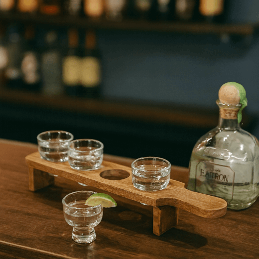 Tequila bottle and shot glasses on a wooden tray with a bar setting in the background.
