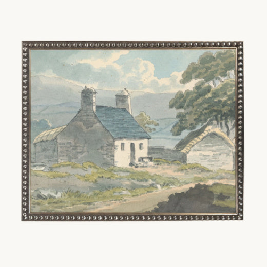 A framed watercolor landscape painting depicting a stone cottage with two chimneys, a thatched outbuilding, trees, and distant hills under a cloudy sky, set in a beaded frame.