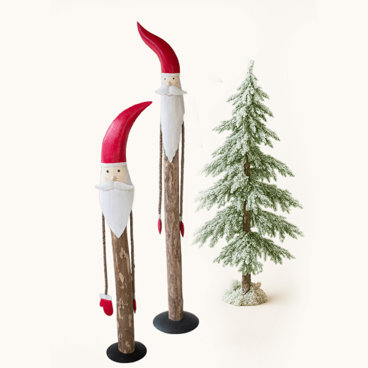 Two tall, whimsical wooden Santa figurines with red hats, white beards, and rope arms standing beside a snow-covered Christmas tree on a light background.