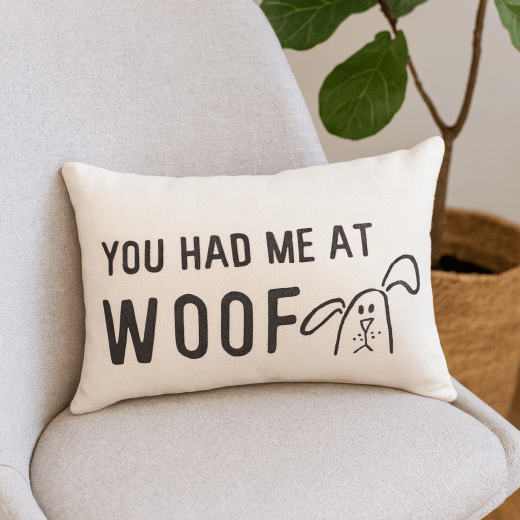 Cream-colored lumbar pillow on a neutral chair featuring the text “You Had Me at Woof” with a hand-drawn dog illustration.