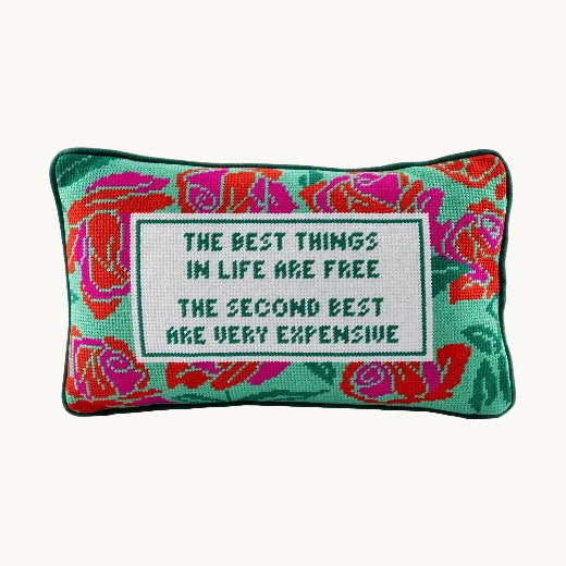 A hand-stitched needlepoint accent pillow featuring a colorful rose design and bold typography rests on a neutral velvet upholstered bench seat by a bright window with a potted fern.