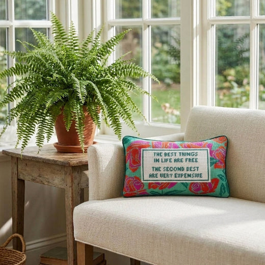 A hand-stitched needlepoint accent pillow featuring a colorful rose design and bold typography rests on a neutral velvet upholstered bench seat by a bright window with a potted fern.