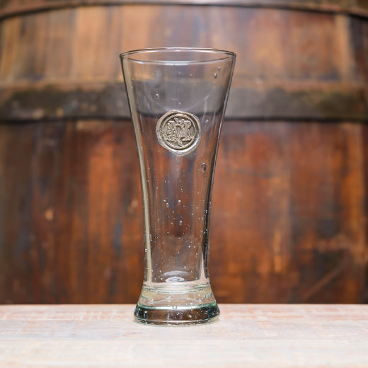 A clear pilsner glass with a pewter initial 'M' emblem, placed on a wooden surface with a barrel in the background.