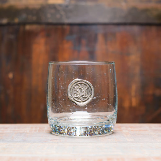 A clear double old fashioned glass with a pewter initial 'M' on the front, displayed against a wooden backdrop.
