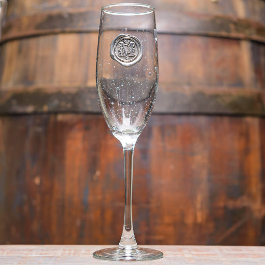 A clear glass flute with a pewter initial 'M' logo on the front, displayed against a wooden backdrop.