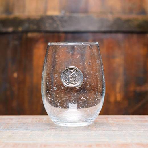 A clear stemless wine glass with a pewter initial embossed on the front, displayed with a wooden background