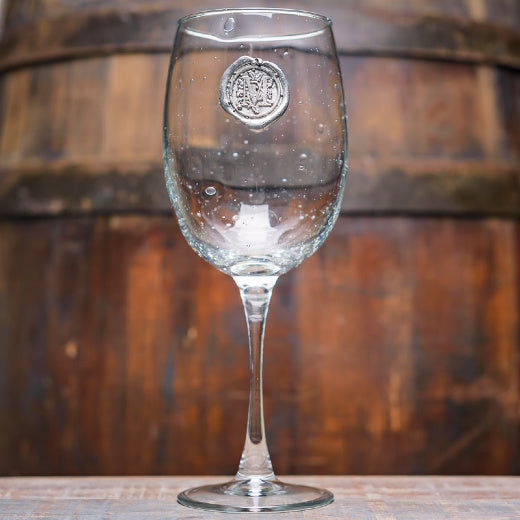 A clear wine glass with a pewter initial emblem on the stem, displayed against a wooden backdrop.